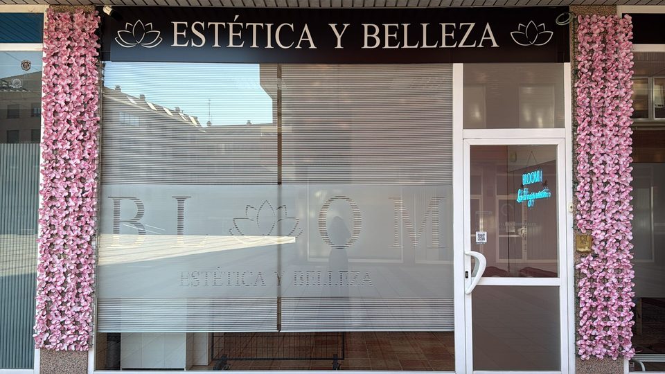 Fachada de Bloom Estética y Belleza en la Avenida Alcalde Miguel Castaño 18 de León: rótulo marrón oscuro con el logo de la flor de loto y el letrero «Estética y Belleza», escaparate con el nombre grabado y dos columnas de flores rosas a los lados de la puerta