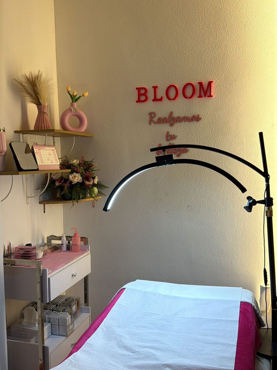 Cabina de pestañas de Bloom: la camilla con la manta fucsia, la lámpara de arco, el carro con las bandejas de pestañas y la pared con el rótulo BLOOM y el lema «Realzamos tu belleza»